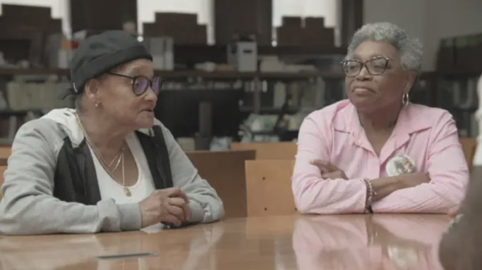 Two older women sit at a table with boxes behind them, looking toward each other.